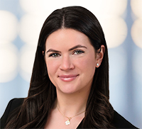 photo with long dark hair, wearing a black blazer, small earrings, and a necklace, poses in front of a blurred light blue background.