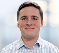 A young man with short brown hair wearing a light plaid shirt stands in front of a softly blurred background.