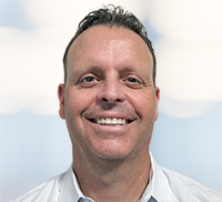 Man with short dark hair smiling at the camera, wearing a light-colored collared shirt, with a blurred light background.