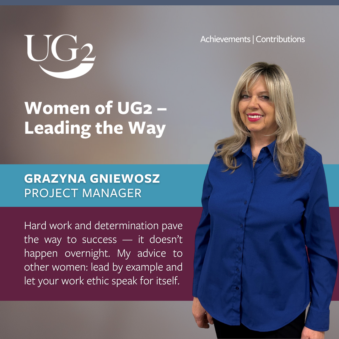 Project manager Grazyna Gniewosz stands smiling in front of a UG2 banner titled "Women of UG2—Leading the Way," sharing her advice on hard work and determination as she inspires others by leading the way.