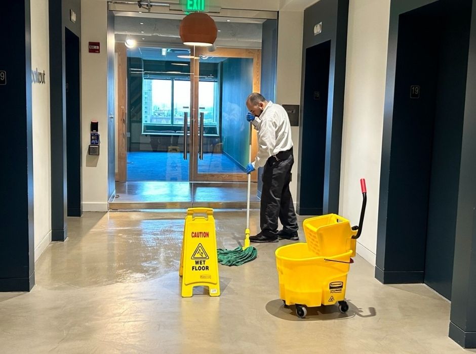 floorcare-janitorial02 A person mops the floor in a hallway with a "Caution Wet Floor" sign and a yellow mop bucket nearby, demonstrating safe janitorial services practices.