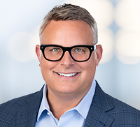 Smiling Midwest man with short gray hair, black-rimmed glasses, and a blue checked blazer over a light blue shirt, standing in front of a blurred light background.