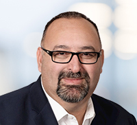 A man with short dark hair, glasses, and a goatee, wearing a white shirt and dark blazer, poses for a professional headshot against a blurred light background, capturing a polished Southern California style.