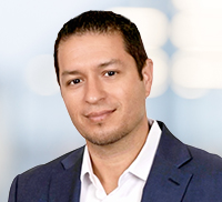Man with short dark hair wearing a white shirt and dark blazer, posing and smiling slightly in front of a blurred Southern California background.