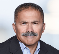 A middle-aged man with short hair and a mustache, wearing a dark blazer and light blue shirt, poses for a formal portrait against a blurred background, evoking the professional style often seen in Southern California.