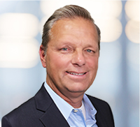 A middle-aged man with short, light hair wearing a dark suit jacket and light blue shirt, standing against a softly blurred Southern California background.