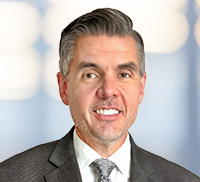A man in a suit and tie smiles at the camera in front of a blurred background with soft lighting, capturing the approachable charm often seen in northern California professionals.