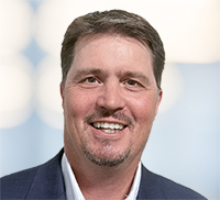 Smiling man with short brown hair and a goatee, wearing a dark suit jacket and light shirt, standing in front of a blurred background with round light patterns, evoking the vibrant energy of Northern California.