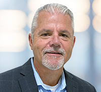 A middle-aged man with short gray hair, a trimmed goatee, and a mustache, wearing a blue shirt and dark blazer, posed against a blurred light background in Southern California.