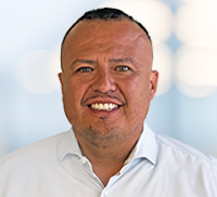 A man with short dark hair and a trimmed beard smiles at the camera, wearing a white collared shirt against a blurred light background, capturing the relaxed vibe of Southern California.