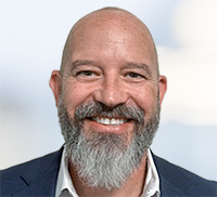 Bald man with a gray beard and mustache, wearing a suit jacket and white shirt, smiling at the camera against a blurred light background, exuding the friendly charm often found in Northern California.