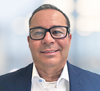 A man wearing glasses, a white shirt, and a dark blazer is smiling at the camera with a blurred Southern California background.