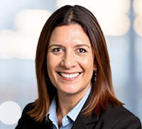 A woman with straight brown hair, wearing a black blazer and light blue shirt, smiles at the camera against a blurred Southern California background.