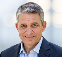 A middle-aged man with short gray hair wearing a dark suit and light blue shirt, smiling at the camera against a blurred light Southern California background.