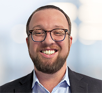 Man with glasses, beard, and short hair wearing a suit jacket and checkered shirt, smiling in front of a blurred light background in Northern California.
