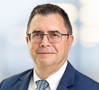 Man wearing glasses, a navy suit, white shirt, and patterned tie, representing mid-atlantic integrated facility services, poses confidently against a blurred light blue background.
