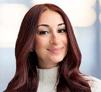 Woman with long, wavy auburn hair and a light sweater smiling at the camera, radiating warmth against a blurred light background—capturing the friendly spirit of the Tri-State Area.