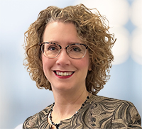 Smiling woman with curly light brown hair, wearing glasses, patterned top, and necklace, posing in front of a softly blurred background.