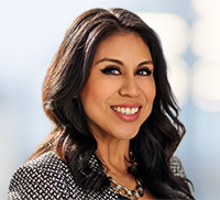 Smiling woman with long dark hair wearing a patterned blazer and necklace, pictured against a blurred light blue background that evokes the relaxed vibe of Southern California.