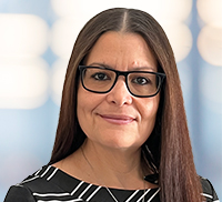A woman with long straight brown hair and glasses, wearing a black and white patterned top, stands in front of a blurred background with blue and white tones in the Tri-State Area.