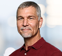 Middle-aged man with short gray hair and trimmed beard, wearing a maroon polo shirt, standing in front of a blurred light background that evokes the relaxed atmosphere of Northern California.