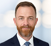 A man with short brown hair, a trimmed beard, and mustache wearing a navy suit, white shirt, and tie poses confidently in front of a blurred light background, embodying the professional style of the Tri-State Area.