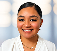 A woman with dark hair pulled back, wearing hoop earrings, a necklace, and a white top, smiles at the camera against a blurred light background reminiscent of a bright New England morning.