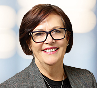 Woman with short brown hair and glasses, wearing a gray blazer and black top, smiling at the camera with a blurred light background, evoking the relaxed professionalism often found in Northern California.