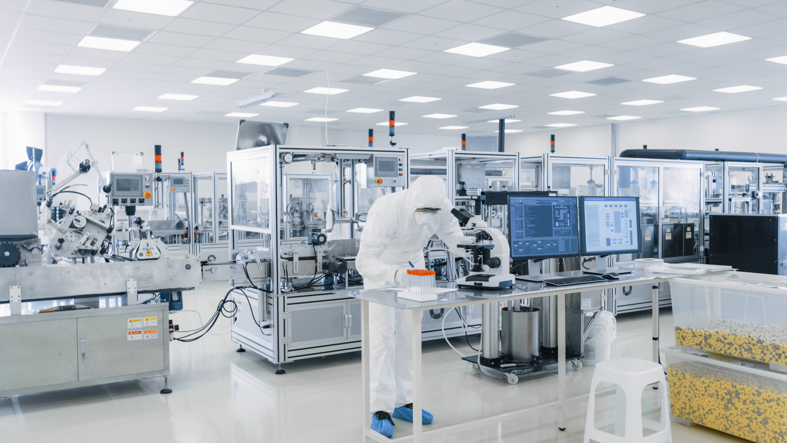 A person in protective clothing works at a computer and microscope in a clean, modern laboratory with automated equipment and bright lighting.