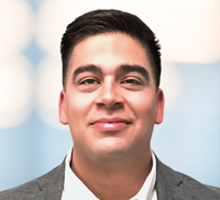 A man with short dark hair, wearing a gray suit jacket and white shirt, smiles at the camera against a blurred light blue and white background, representing Midwest Integrated Facility Services with professional confidence.