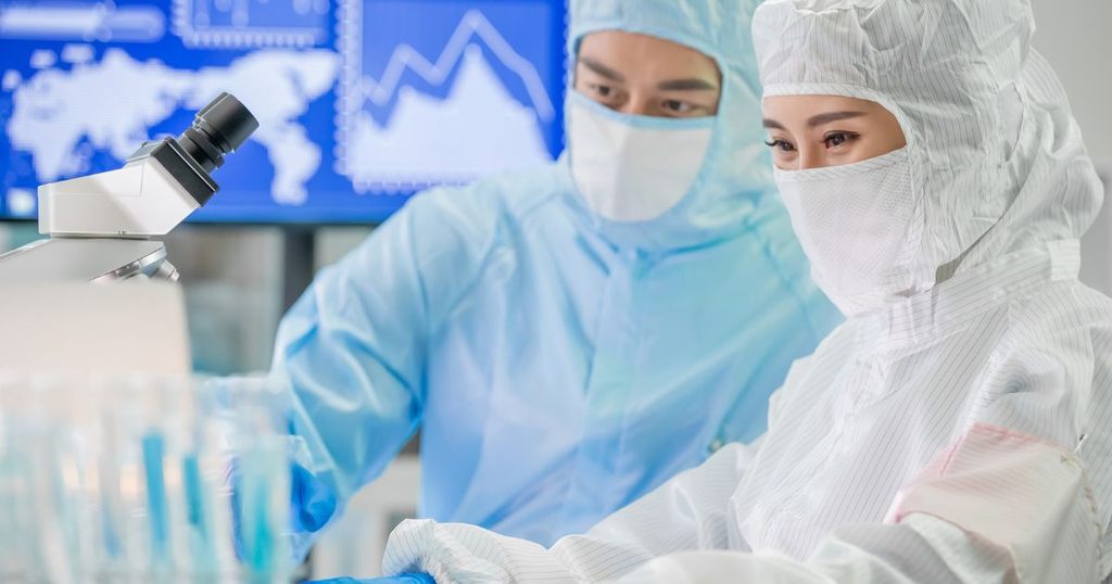 Two scientists in protective suits work in a laboratory, which benefits from professional cleanroom facility services, with one using a microscope and data charts visible on a screen in the background.