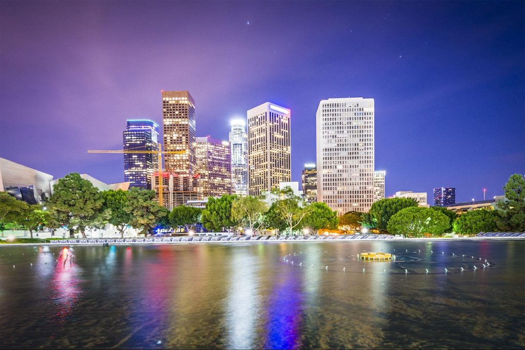 Los Angeles skyline at night reflecting in a park pond with trees in the foreground.